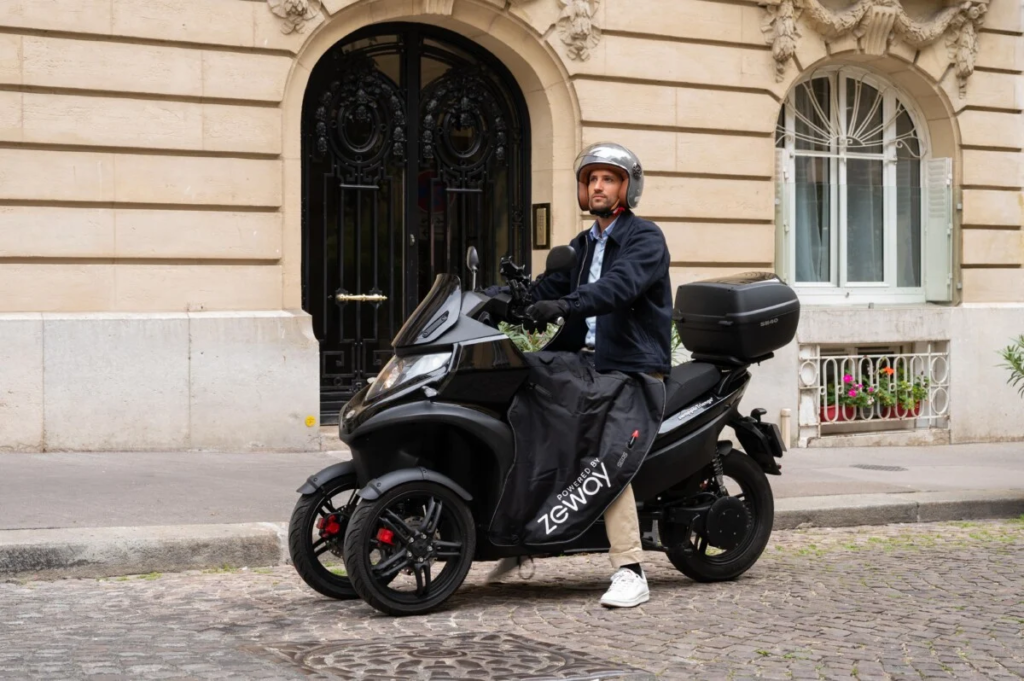 Homem pilotando um triciclo elétrico, ele esta com capacete e olhar fixado para frente, o triciclo esta com baú na garupa.